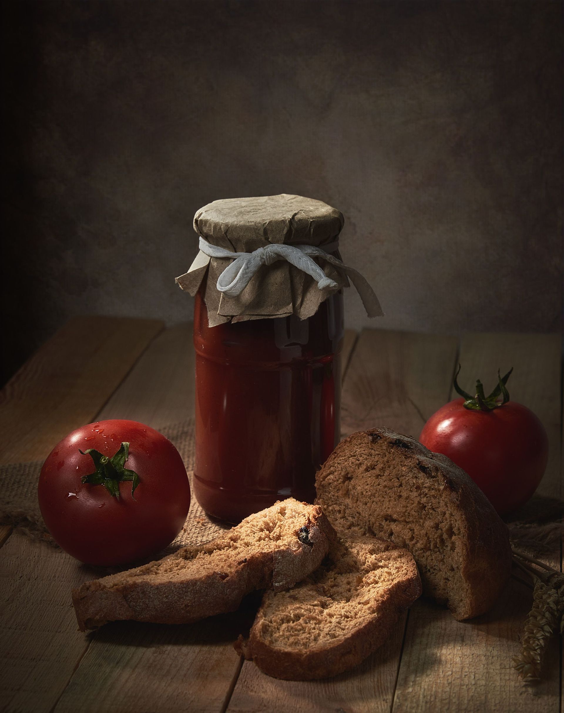 A rustic still life featuring tomatoes, sliced bread, and a jar of sauce on a wooden table.